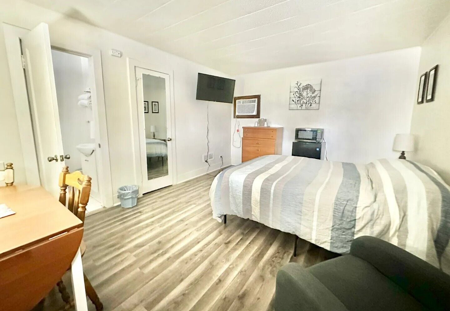 A hotel room with a wooden floor, a bed with striped bedding, a desk with a chair, a television mounted on the wall, and a small cabinet.