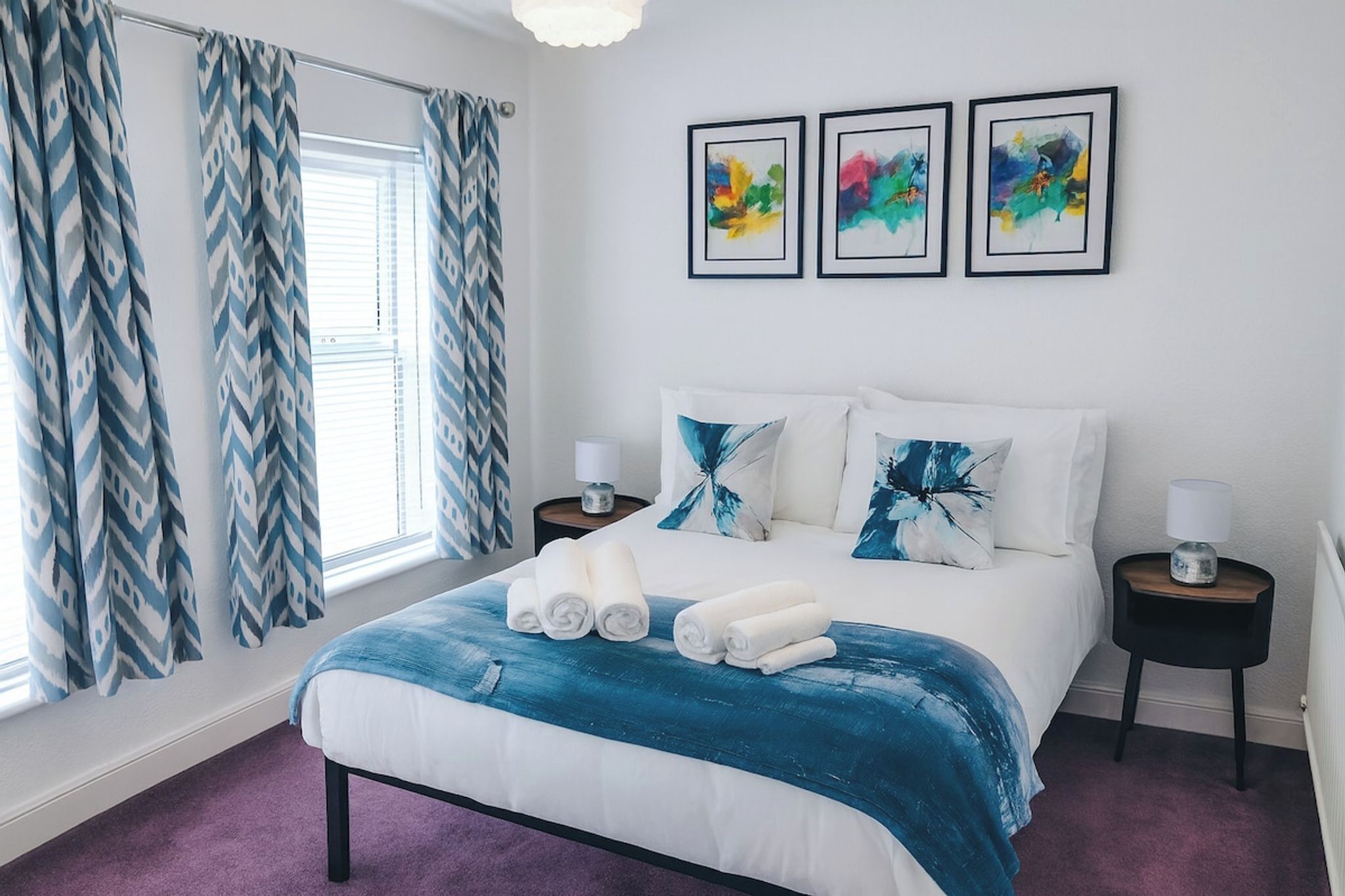 Een slaapkamer met een bed, kussens, een blauwe deken, een raam met gordijnen, en drie kunstwerken in lijsten aan de muur.