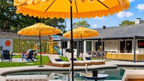Un espace piscine avec des chaises longues et des parasols, une aire de jeux pour enfants et un bâtiment orné d’un panneau Netflix.