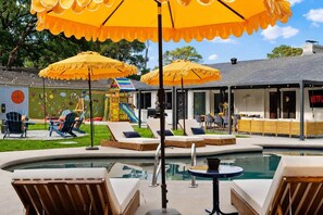 A pool area with lounge chairs and umbrellas, a children's play structure, and a building with a Netflix sign. - Kaleo - Pickleball | Pool | Hot Tub | Central Spot (Irving)