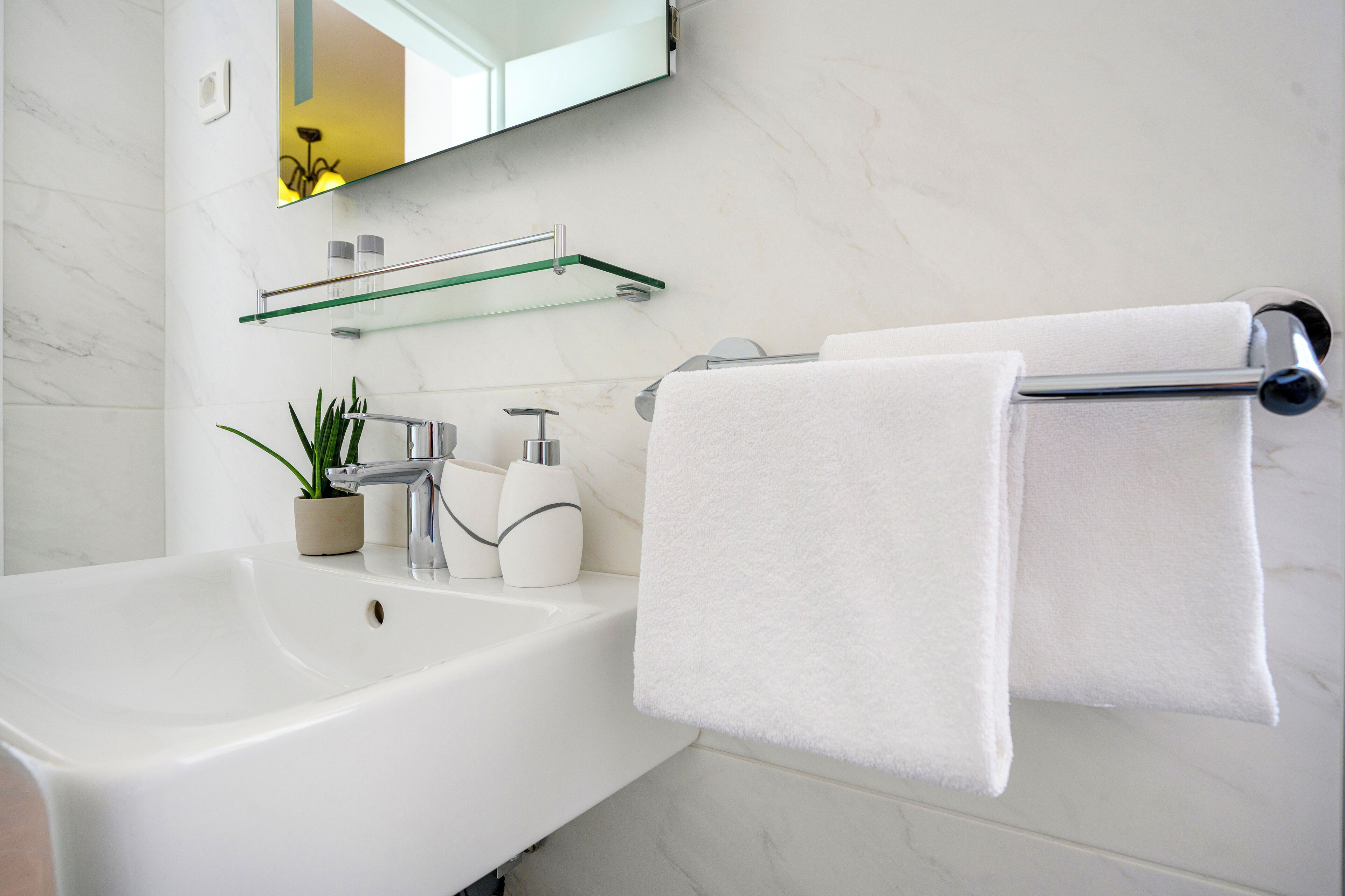 A bathroom with a white sink, a mirror, a towel rack with two towels, a glass shelf with toiletries, and a potted plant.