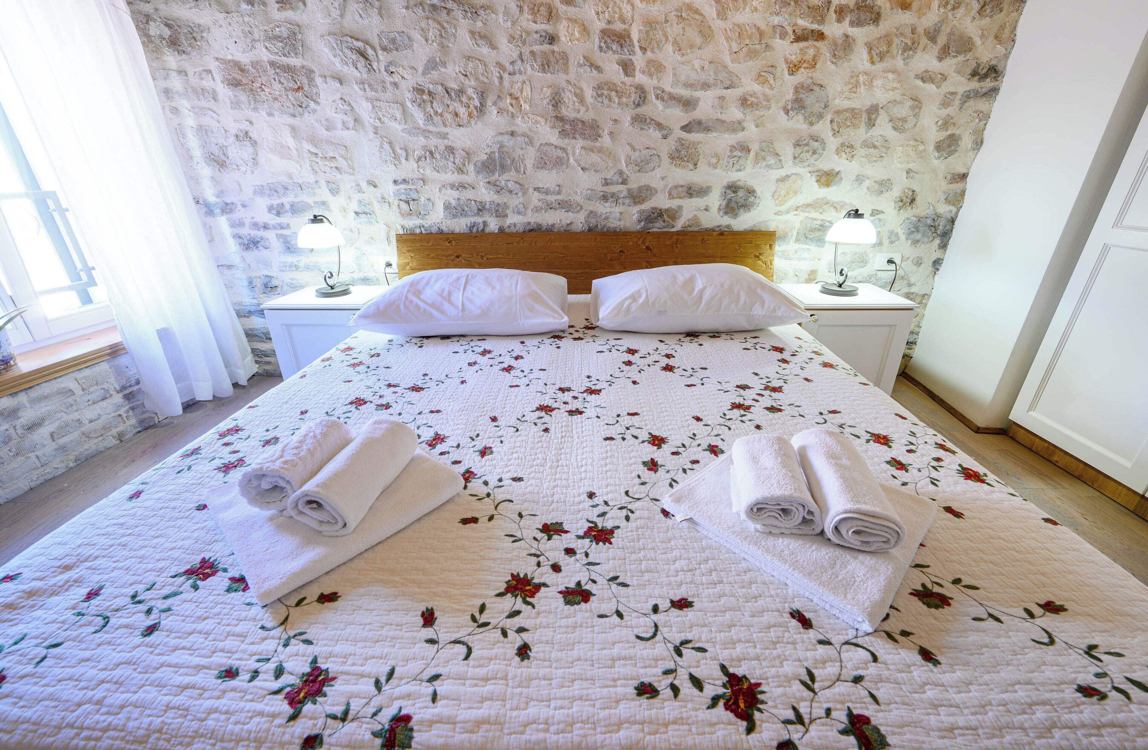 A bedroom with a stone wall, a wooden headboard, two white pillows, a floral quilt, and two rolled towels on the bed.