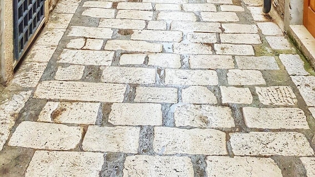 Una estrecha calle empedrada flanqueada por edificios históricos de piedra con contraventanas y un farol.