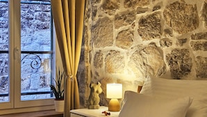 Un dormitorio con pared de piedra, una cama con ropa de cama blanca, una mesita de noche con lámpara y una ventana con cortinas.
