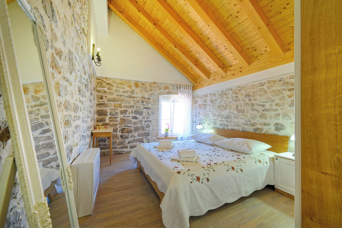 Une chambre avec des murs en pierre, des poutres en bois au plafond, un lit avec du linge de lit blanc, une table de chevet et une fenêtre avec des rideaux.