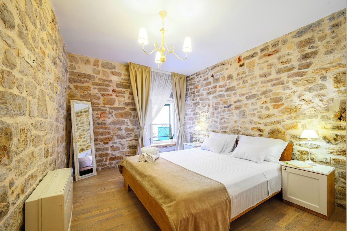 Une chambre avec un mur en pierre, un lit, une table de chevet, un miroir et une fenêtre avec des rideaux.