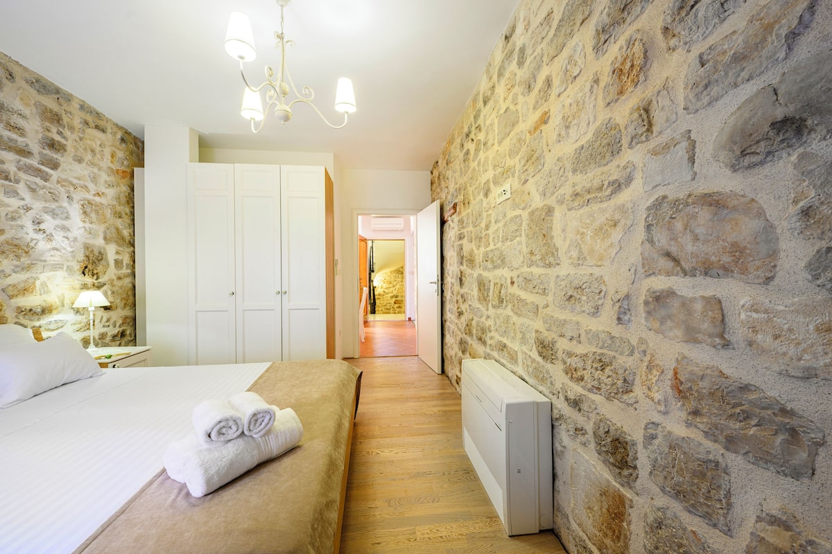 Une chambre avec des murs en pierre, un lit recouvert de draps blancs, une table de chevet avec une lampe, et un couloir menant à une autre pièce.