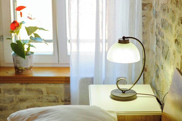 A room with a bed, a bedside table with a lamp, a potted plant, and a window with curtains. - Apartmani Festum (Šibenik)
