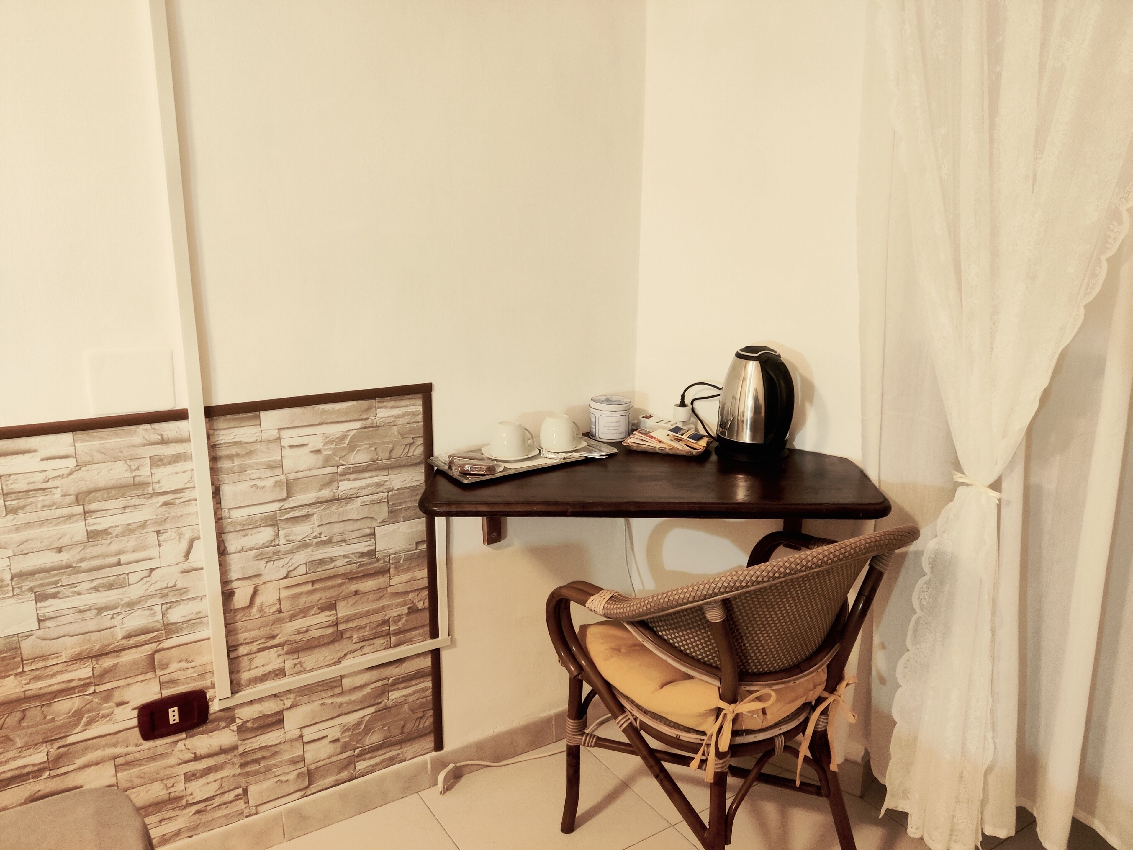 A small room with a stone-patterned wall, a wooden table with a kettle, cups, and newspapers, and a wicker chair with a cushion.