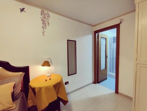 A room with a bed, a table with a lamp, a mirror, and a door leading to another room. - The Second Floor (Naples)