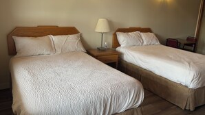 Two single beds with wooden headboards, a bedside table with a lamp, and a small seating area with a chair and table. - Casey's Inn (Lac du Bonnet)