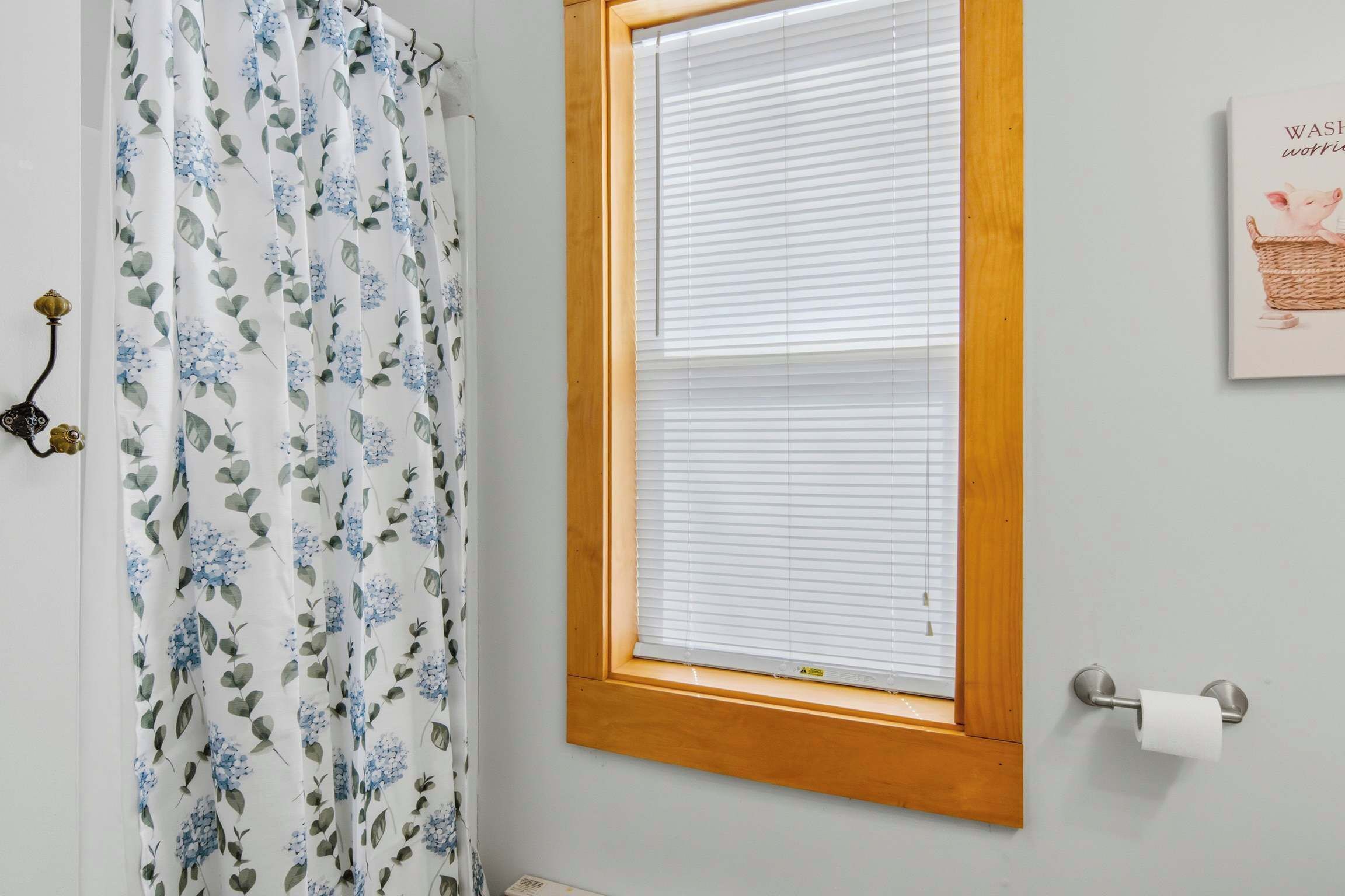 Un baño con cortina de ducha, una ventana con persianas, un dispensador de papel higiénico y un cuadro en la pared.