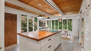 A modern kitchen with a wooden ceiling, white cabinetry, a central island, pendant lighting, and large windows offering views of greenery. - Burke Bay A-Frame Retreat w/Cedar Hot Tub - 15 Minutes to Seattle Ferry (Bremerton)