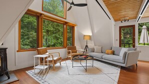A modern living room with a high ceiling, large windows, a ceiling fan, a sofa, a coffee table, and a lamp. - Burke Bay A-Frame Retreat w/Cedar Hot Tub - 15 Minutes to Seattle Ferry (Bremerton)