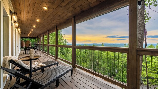 Un terrazzo in legno con posti a sedere all'aperto, vista su un paesaggio boschivo e un tramonto sullo sfondo.