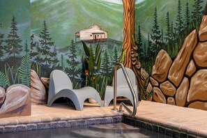 A spa area with a stone wall, a small wooden cabin mural, and a pool. - Indoor Pool, Mtn Views, Hot Tub, Couples Cabin, Fire Pit, & Close to Strip (Pigeon Forge)