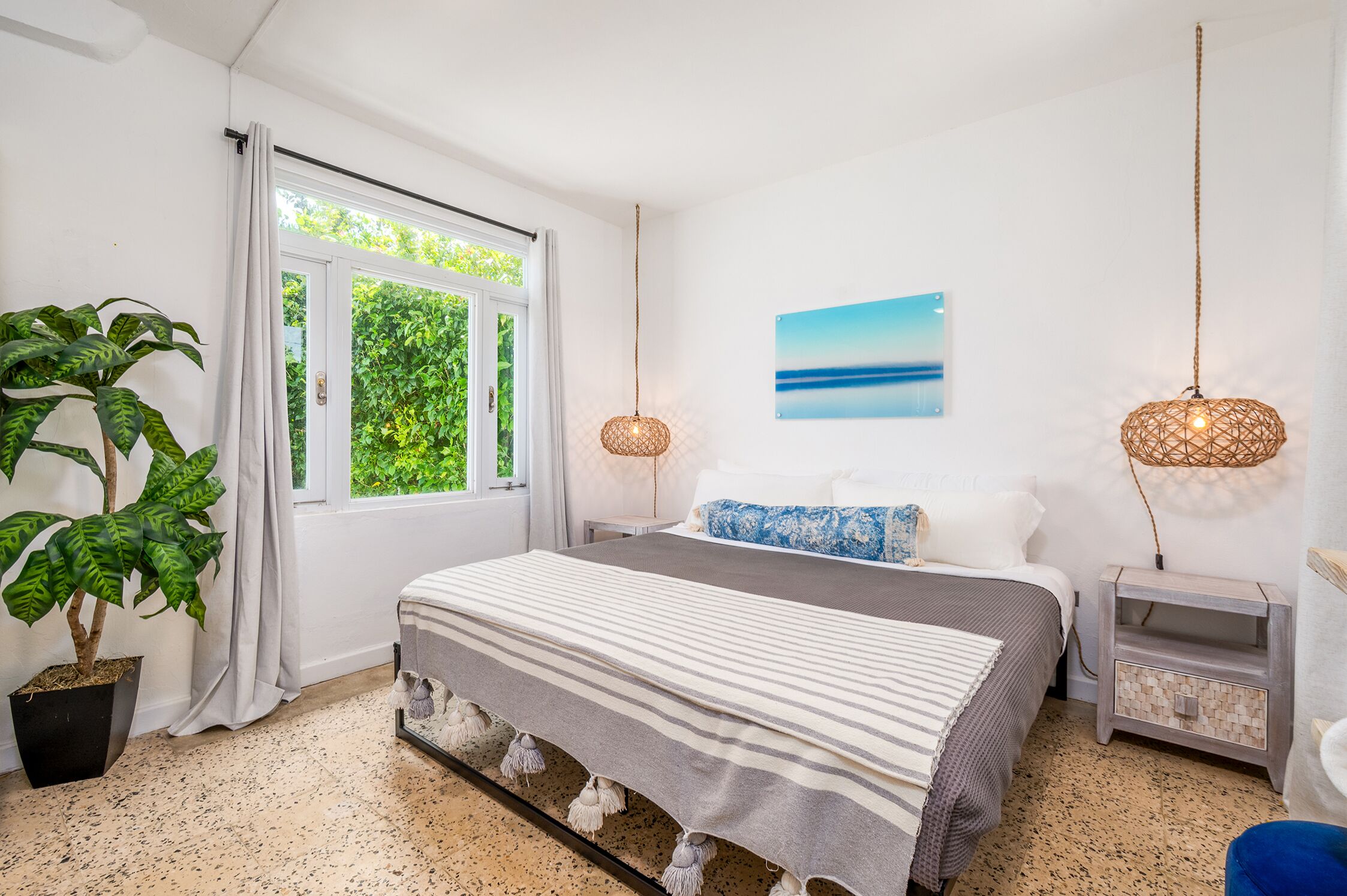 Un dormitorio con cama, planta en maceta, ventana, un cuadro y dos lámparas de noche.