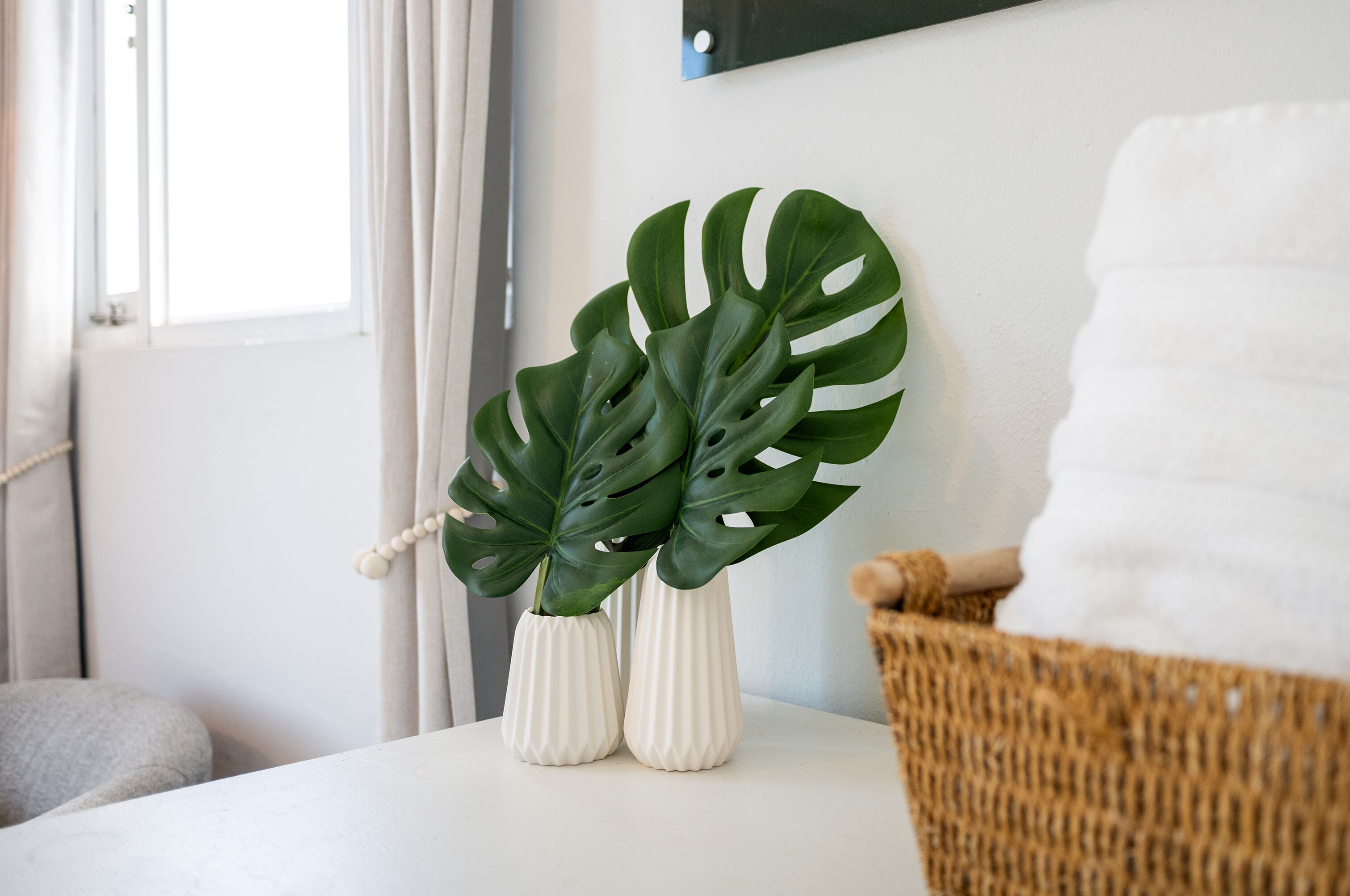 Two white vases with green monstera leaves on a white table, a wicker chair, and a grey cushion in a room with a white wall and a window with curtains.