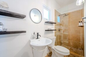 A bathroom with a sink, toilet, and shower. There are shelves with toiletries and a mirror on the wall.