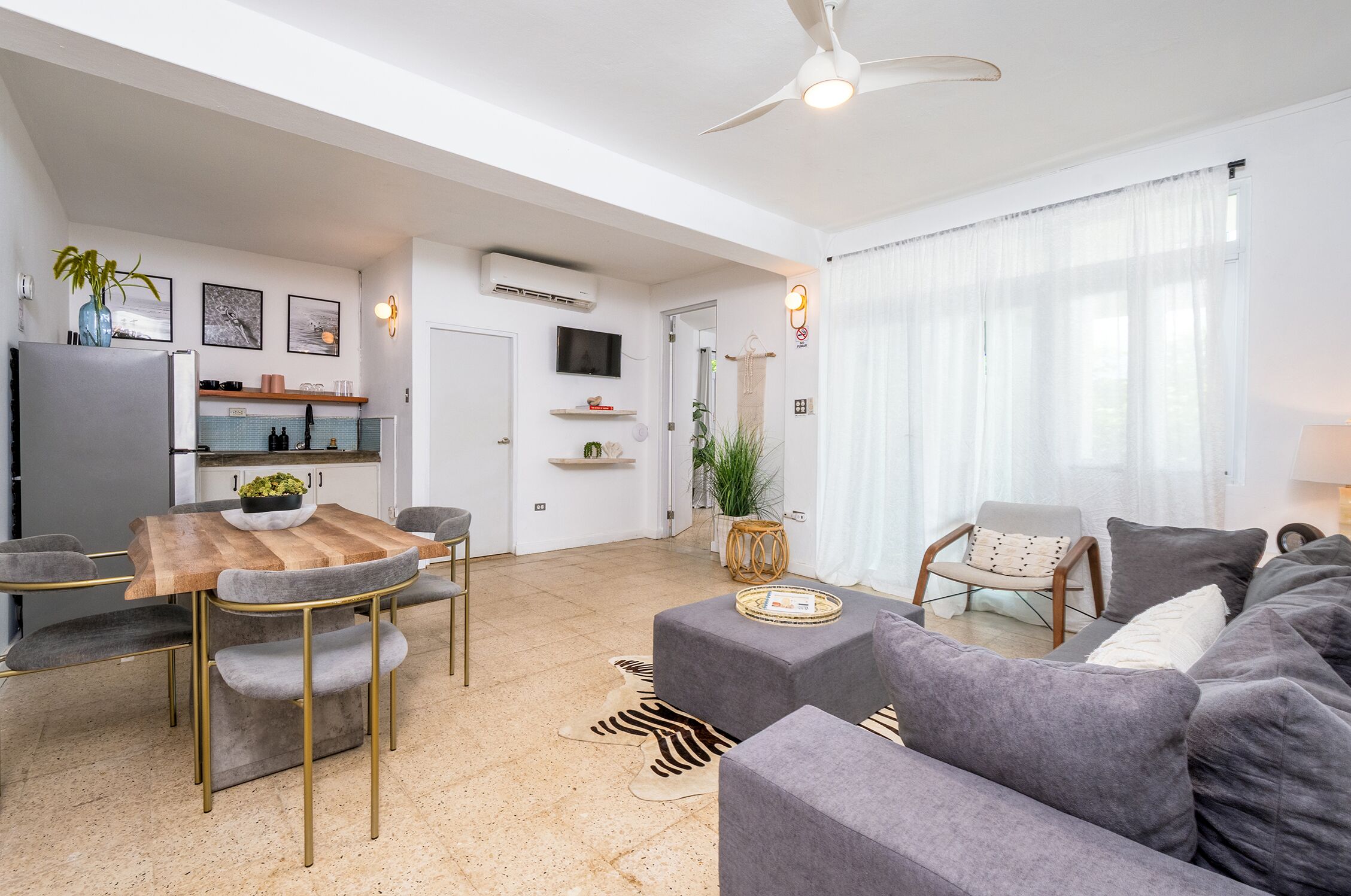 Una sala moderna con un sofá gris, una mesa de comedor de madera con sillas, un ventilador de techo y una zona de cocina con televisión y aire acondicionado.
