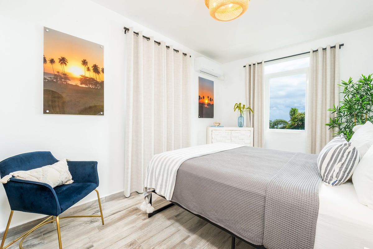 A bedroom with a bed, a chair, a window, curtains, and a wall-mounted air conditioner.