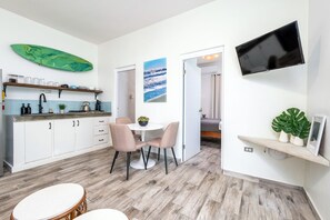 A modern kitchen with white cabinets, a dining area with a round table and chairs, a mounted TV, and a view of the sea through a window.