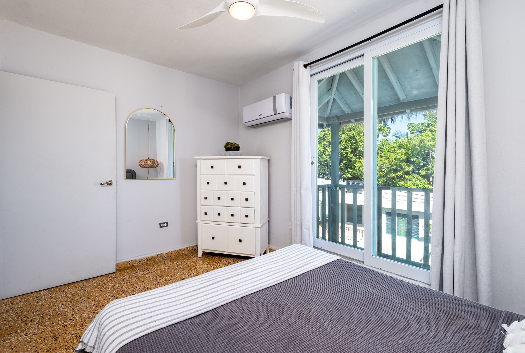 Een slaapkamer met een bed, een dressoir, een spiegel, een airconditioner en een schuifpuien naar een balkon.