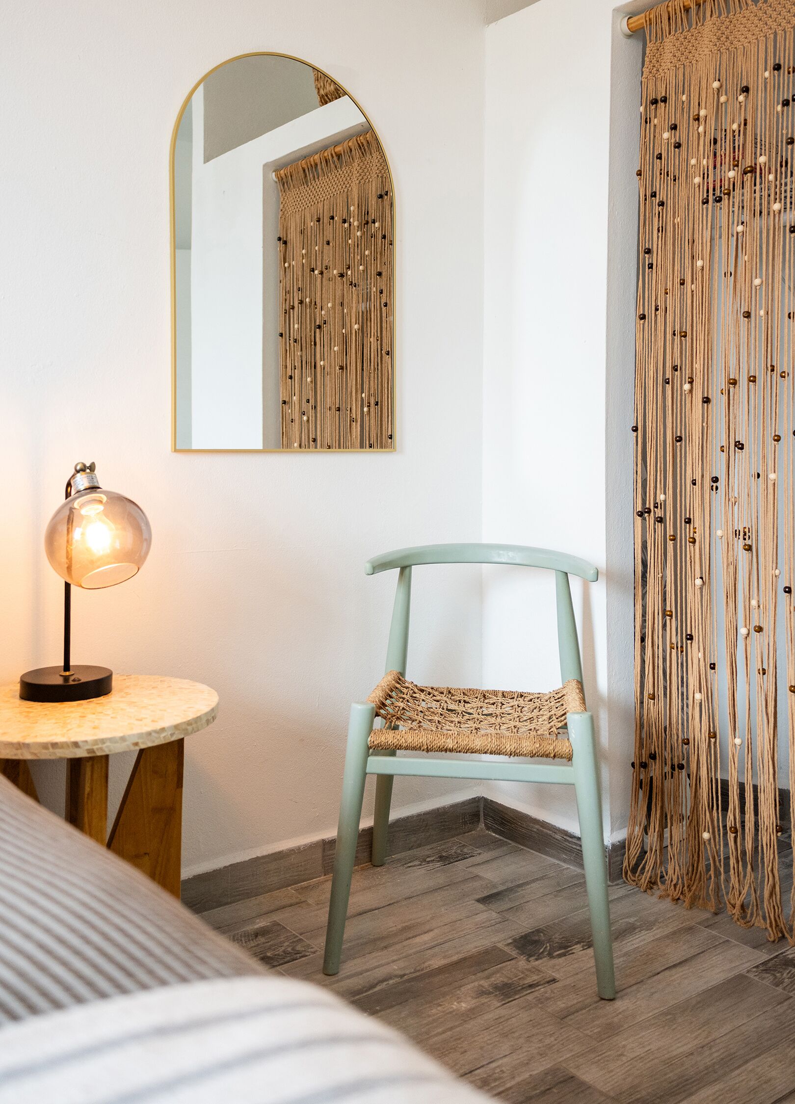 A room with a round table, a lamp, a chair, a mirror, and a macramé curtain.