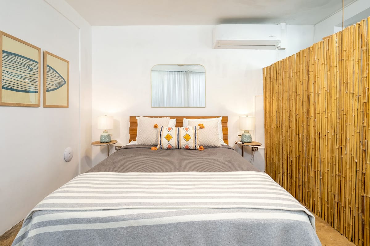 A bedroom with a bed, bedside tables, a window with curtains, and a wall-mounted air conditioner.