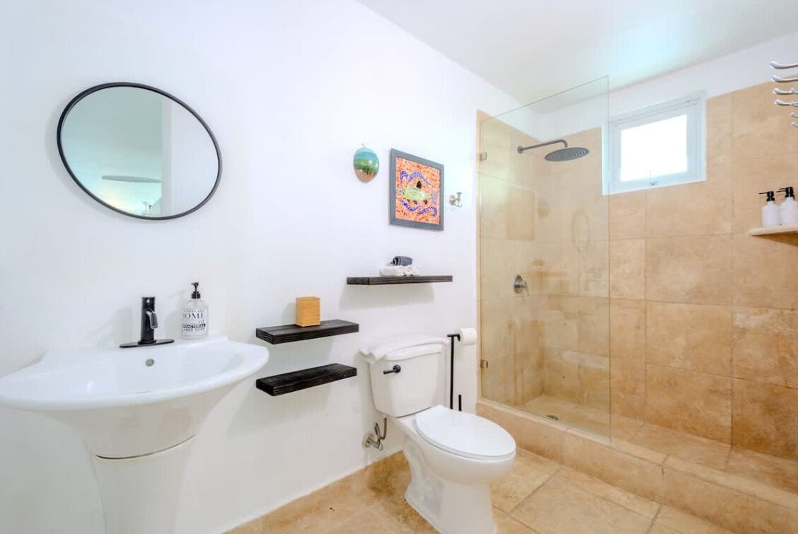 Un baño con lavamanos, inodoro y ducha blancos. Hay repisas con artículos de aseo personal y un cuadro en la pared.