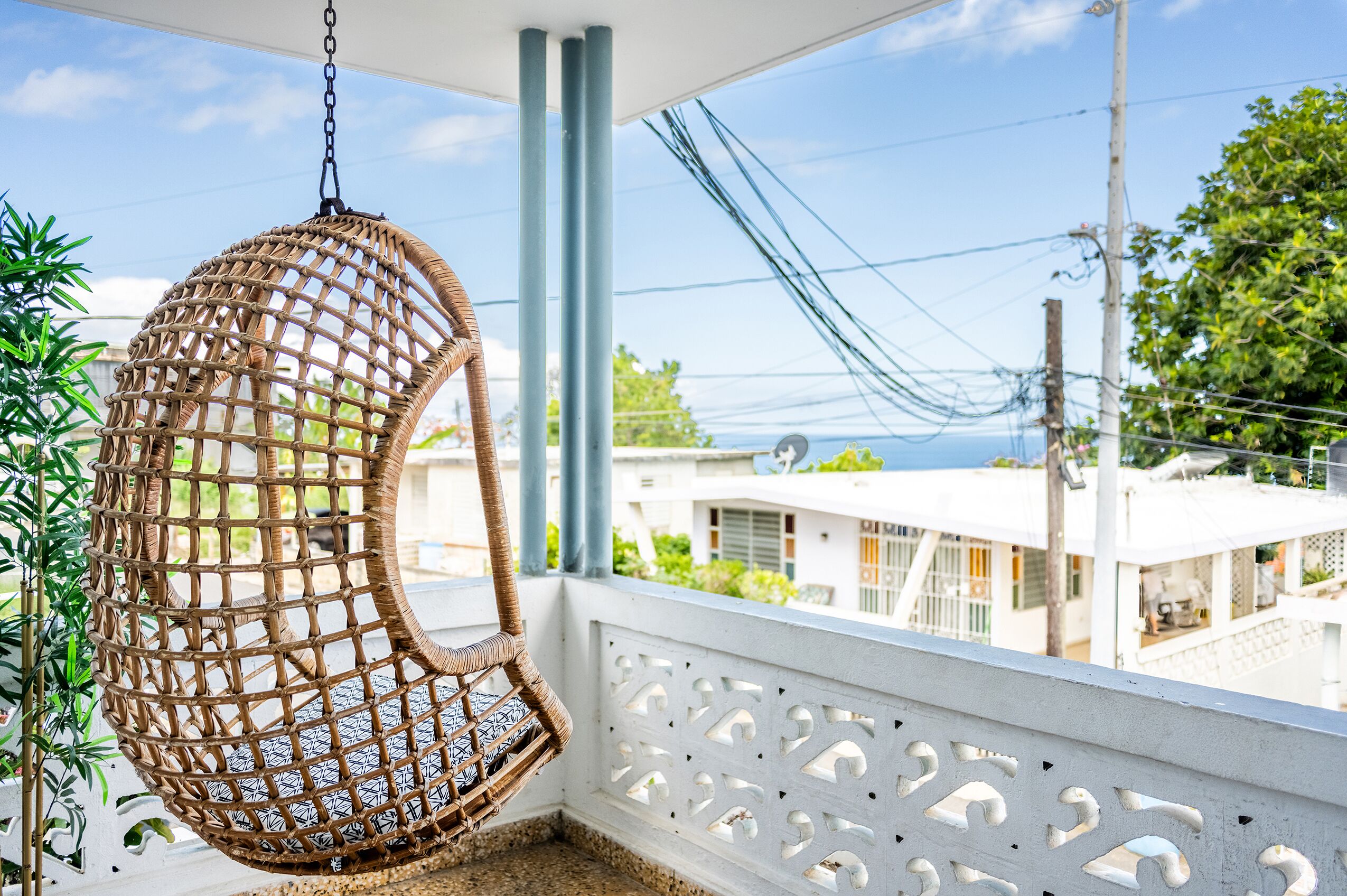 A wicker hanging chair on a balcony with a white railing and a view of buildings and greenery.