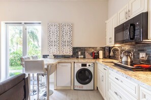 Una cocina moderna con armarios blancos, lavadora integrada, hervidor de acero inoxidable y vistas a zonas verdes.
