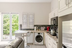 A modern kitchen with a built-in washer and dryer, a large window with a view of greenery, and a bar stool.