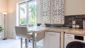 A modern kitchen with a dining area, a dishwasher, a washing machine, and a large window with a view of greenery. - Poolside Boutique Guest House | Fort Lauderdale (Fort Lauderdale)