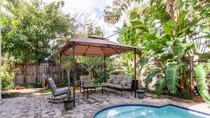 Un patio trasero con piscina, zona de estar techada y abundante vegetación.