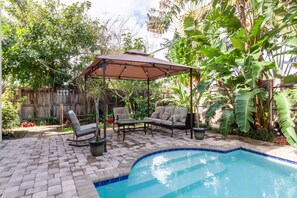 Un patio trasero con piscina, zona de estar techada y abundante vegetación.
