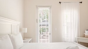 Habitación de hotel con una cama grande, una mesita de noche con lámpara, una puerta que ofrece vistas a zonas verdes y una ventana con cortinas traslúcidas.