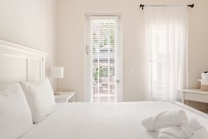 Habitación de hotel con una cama grande, una mesita de noche con lámpara, una puerta que ofrece vistas a zonas verdes y una ventana con cortinas traslúcidas.