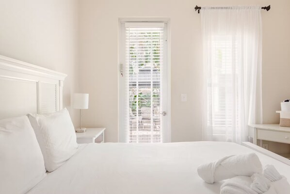 A hotel room with a large bed, a bedside table with a lamp, a door with a view of greenery, and a window with sheer curtains.