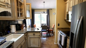 Una cocina con encimeras de granito, refrigerador de acero inoxidable, armarios de madera, un comedor con mesa y sillas, y una ventana con cortinas.