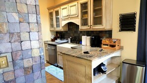 Una cocina con paredes de piedra, armarios de madera, encimera de granito, lavavajillas, cafetera y refrigerador de acero inoxidable.