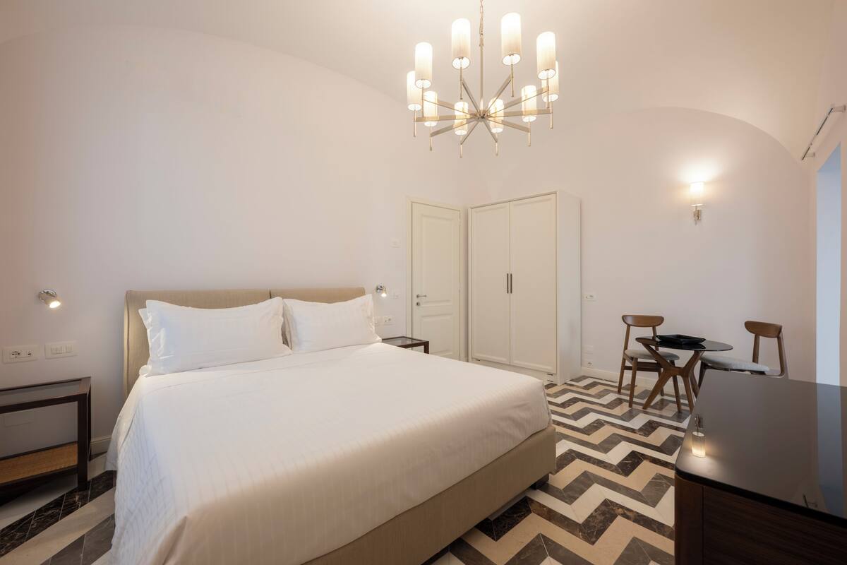 Habitació d'hotel amb un llit gran, una làmpada de cristall, un armari, un escriptori i una cadira.