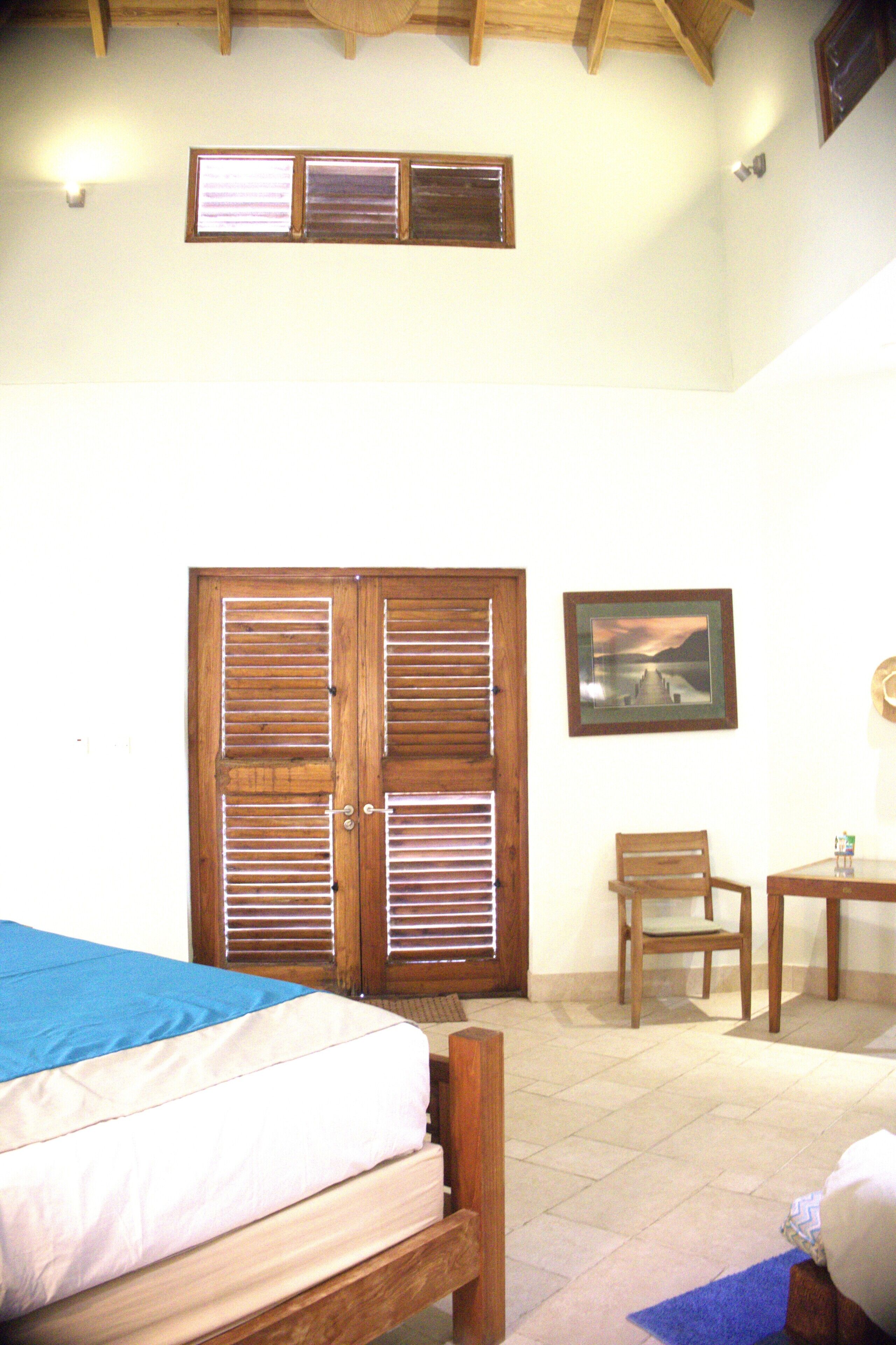 A bedroom with a bed, wooden door with shutters, a chair, a table, and a framed picture on the wall.