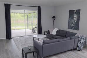 A modern living room with a grey sofa, a small round table, and a large sliding glass door leading to a balcony. - Wesley Chapel Home 3Bed/2Bath with water view (Zephyrhills)
