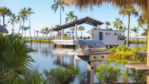 Un barbecue installé près d’un plan d’eau, avec des palmiers et une structure couverte en arrière-plan.