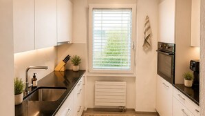 A modern kitchen with white cabinets, a black countertop, a built-in oven, and a window with blinds. - Bern Garten Residence 152m Luxury Apartment (Bern)