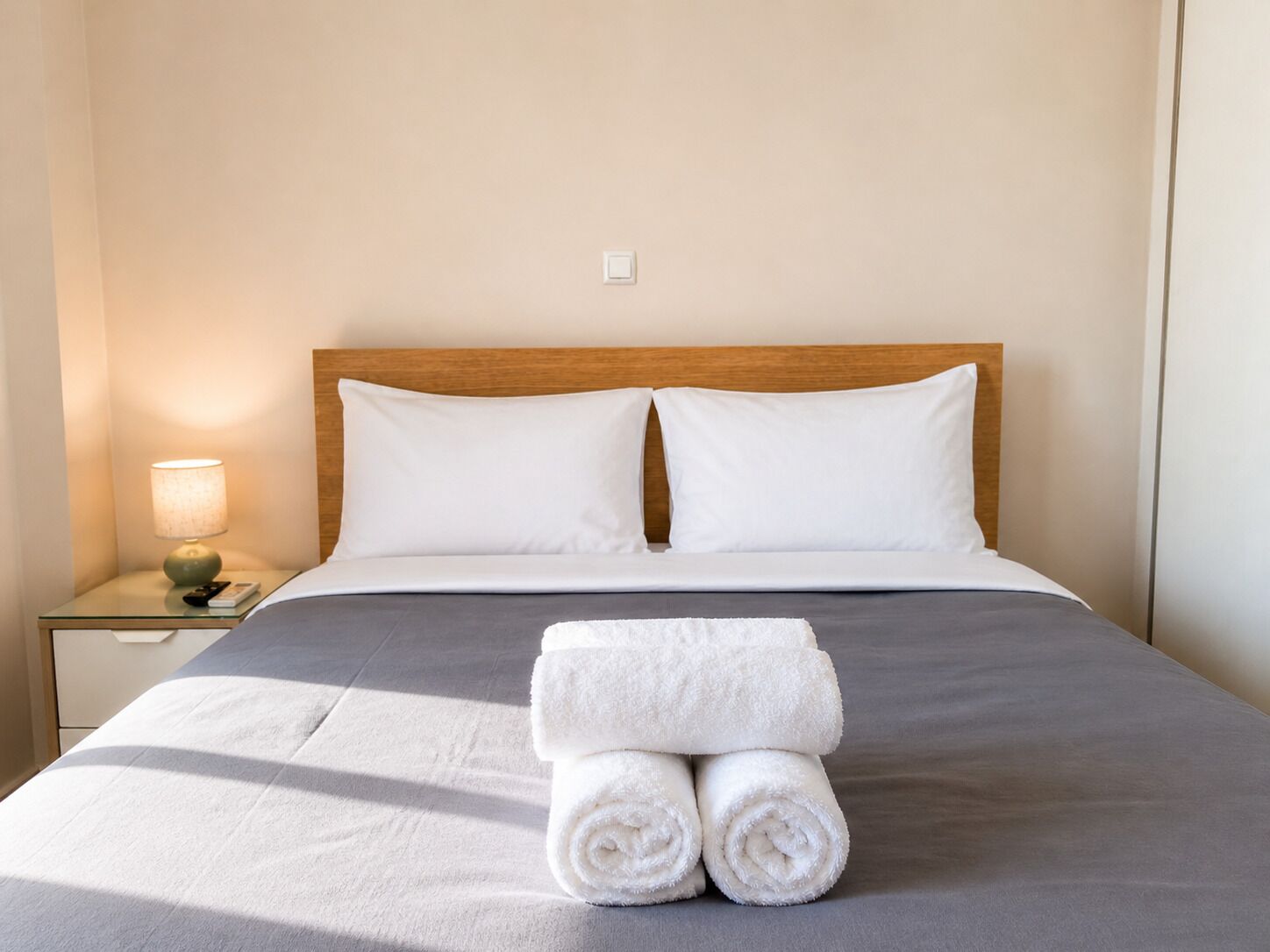 A neatly made bed with white linens and two rolled towels, a wooden headboard, and a bedside lamp on a nightstand.