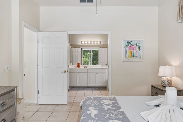 Une chambre à coucher avec un lit, une table de chevet avec une lampe, une salle de bain avec un meuble-lavabo et un miroir, et une porte donnant sur une autre pièce.