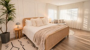 Un dormitorio con una cama grande, mesitas de noche, una planta en maceta y una ventana con cortinas.