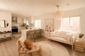 A modern kitchen with white cabinets, a central island with bar stools, a dining area with a sofa and chairs, and large windows providing natural light. - Chic Designer Home, Walkable to Marine Stadium and Minutes to Beach & 2nd Street (Long Beach)
